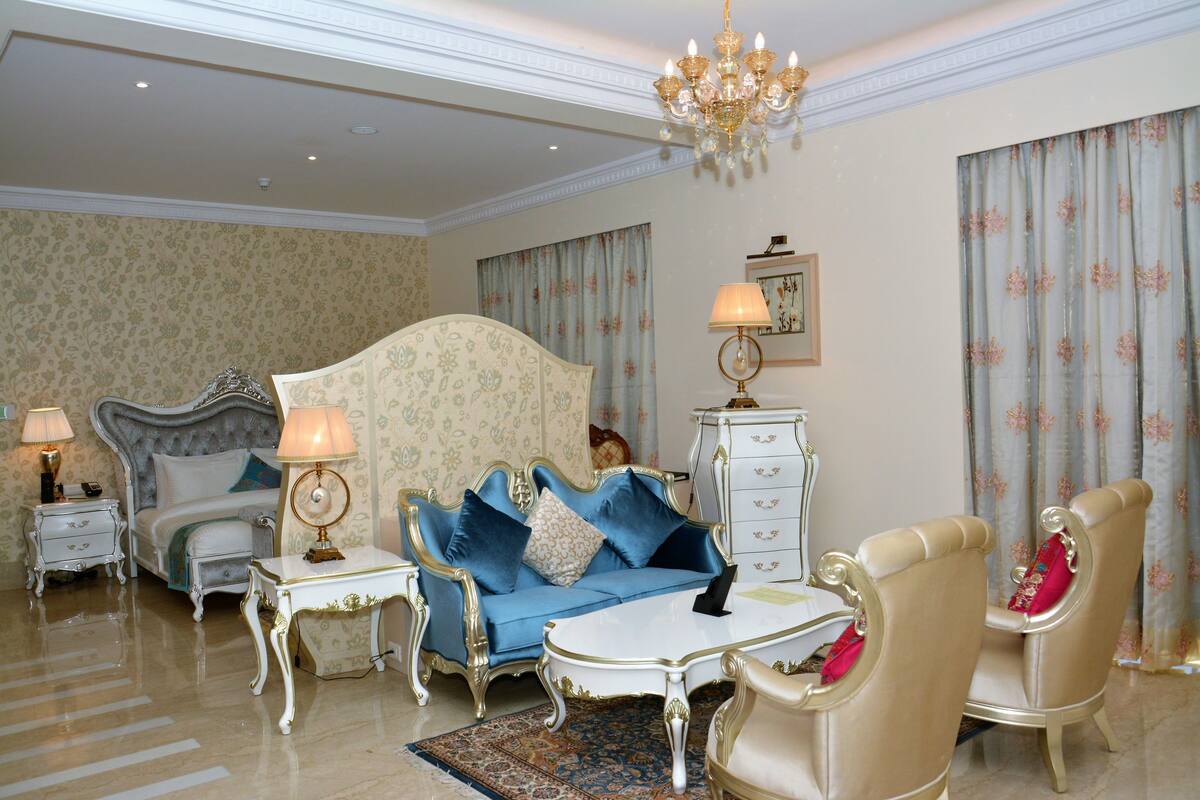 A luxurious bedroom with a large bed, a blue sofa, a white coffee table, and a chandelier.