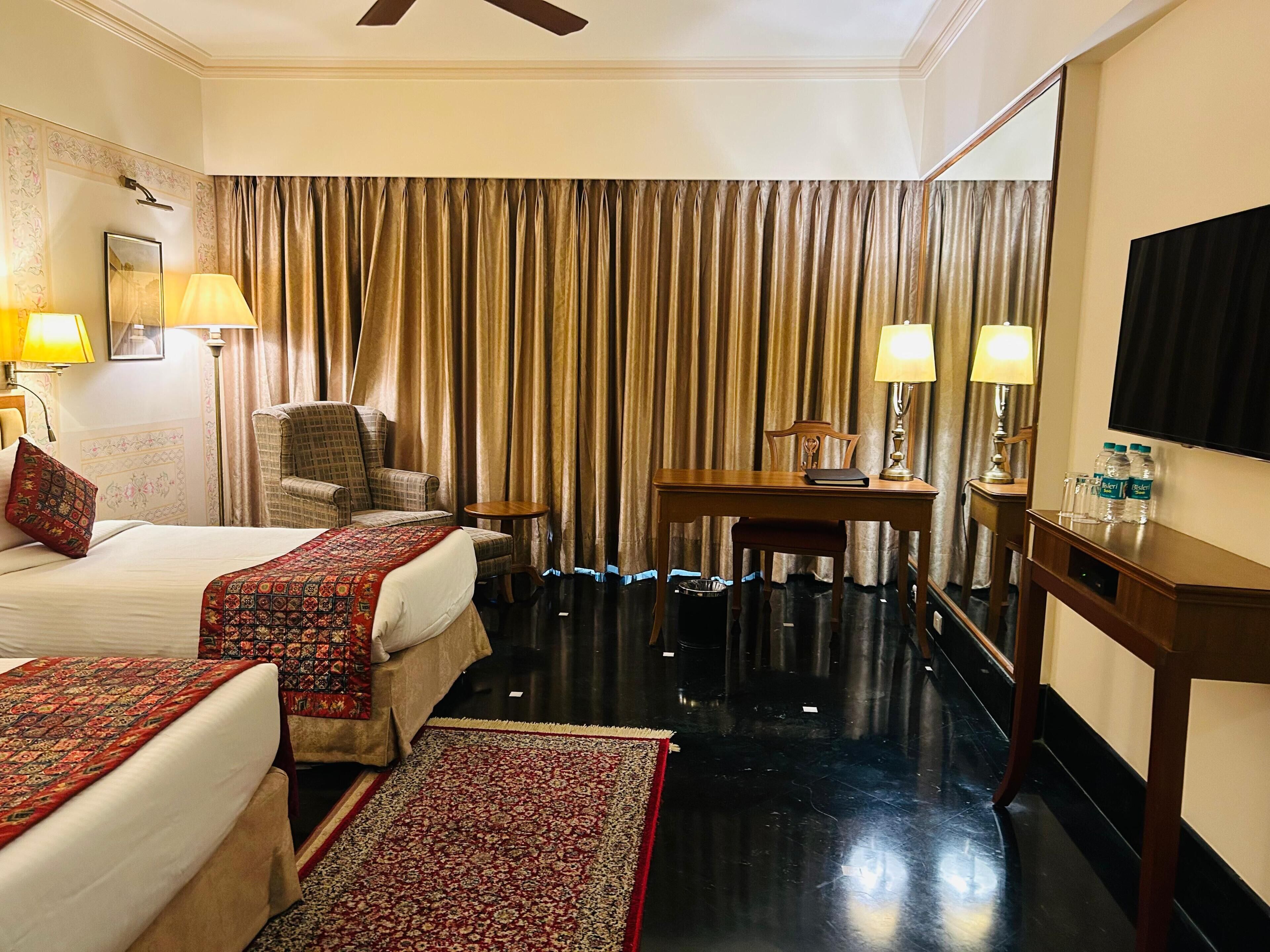 A hotel room with two beds, a desk, a chair, a TV, and a large window with curtains.