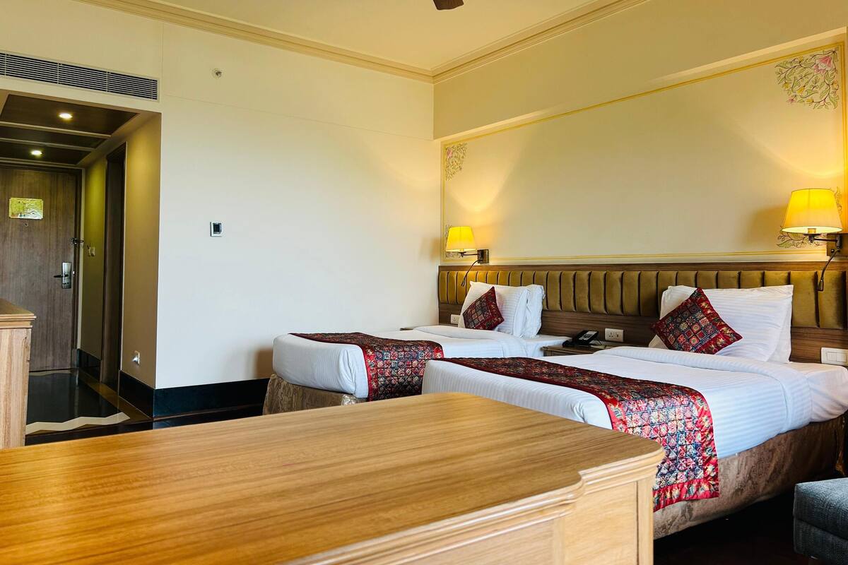 A hotel room with two beds, a wooden desk, a ceiling fan, and a wall-mounted lamp.