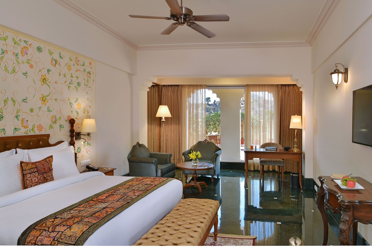 A hotel room with a large bed, a desk, two chairs, a ceiling fan, and a TV on the wall.