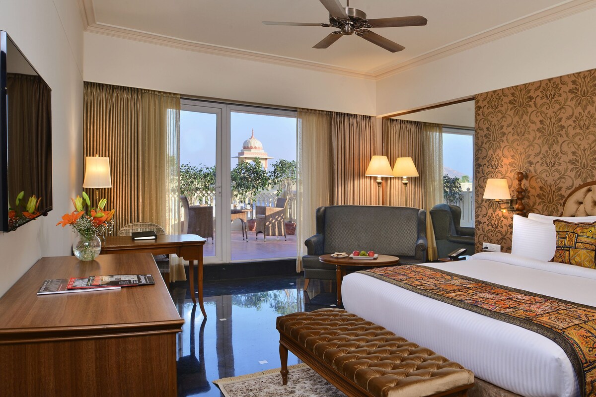A hotel room with a large bed, a desk, a chair, and a view of a building through the window.