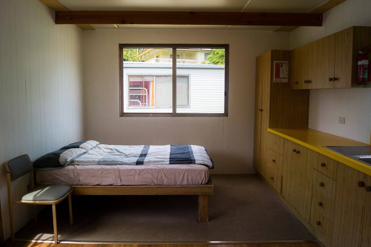 A small, single-bed room with a wooden bed frame, a small chair, a window with a view of a house, and a yellow countertop with cabinets.