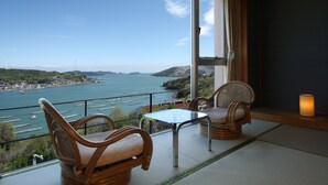 Un balcon avec des fauteuils en osier et une table en verre, offrant une vue sur la côte, avec des bateaux et des collines.