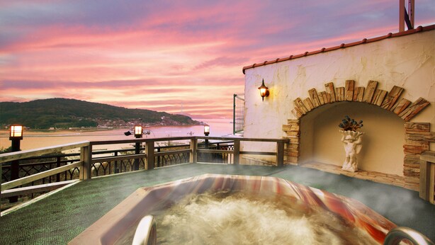 Un bain à remous offrant une vue sur un coucher de soleil se reflétant sur une étendue d’eau, un arc en pierre et une balustrade surplombant le paysage.