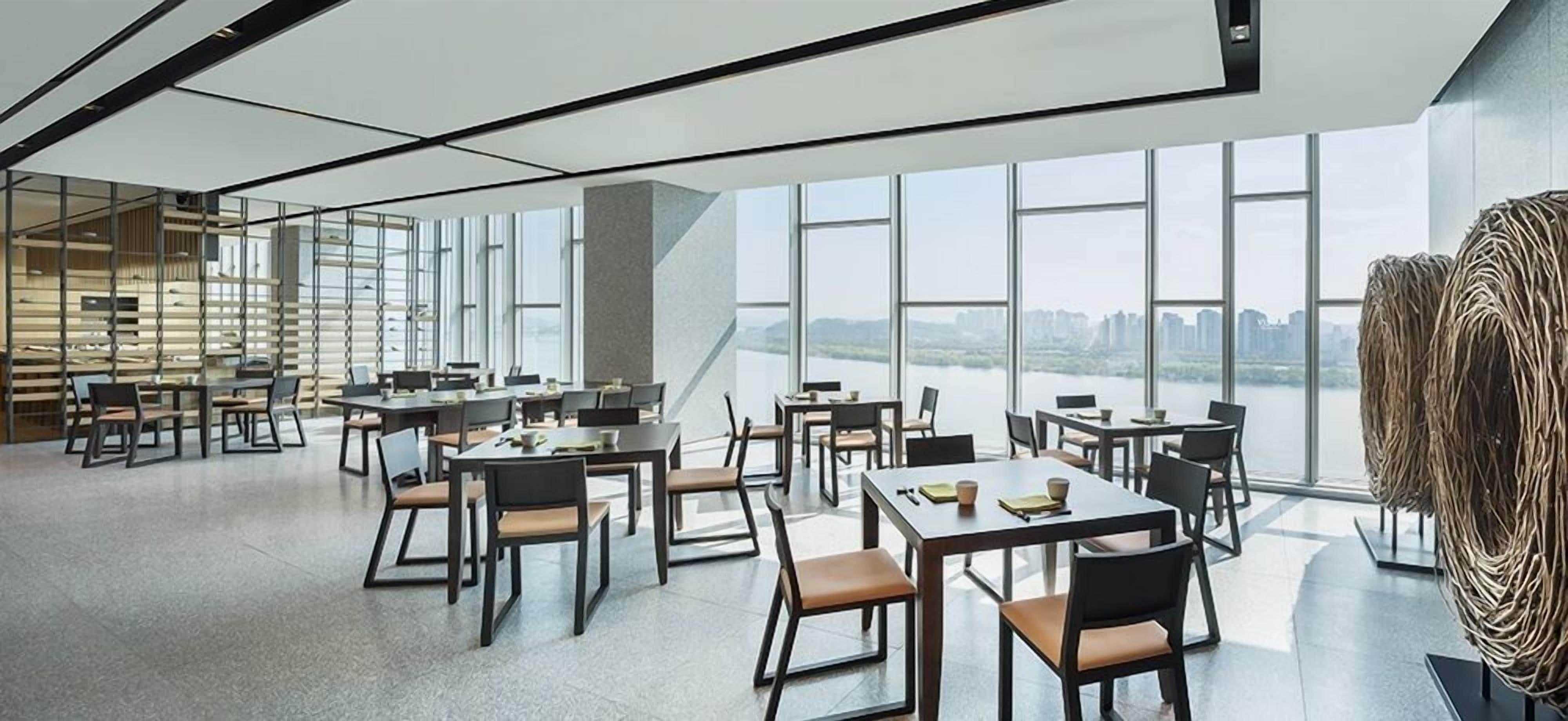 A modern dining area with tables and chairs arranged for groups, large windows offering a view of a river and cityscape, and a unique woven sculpture on the right.