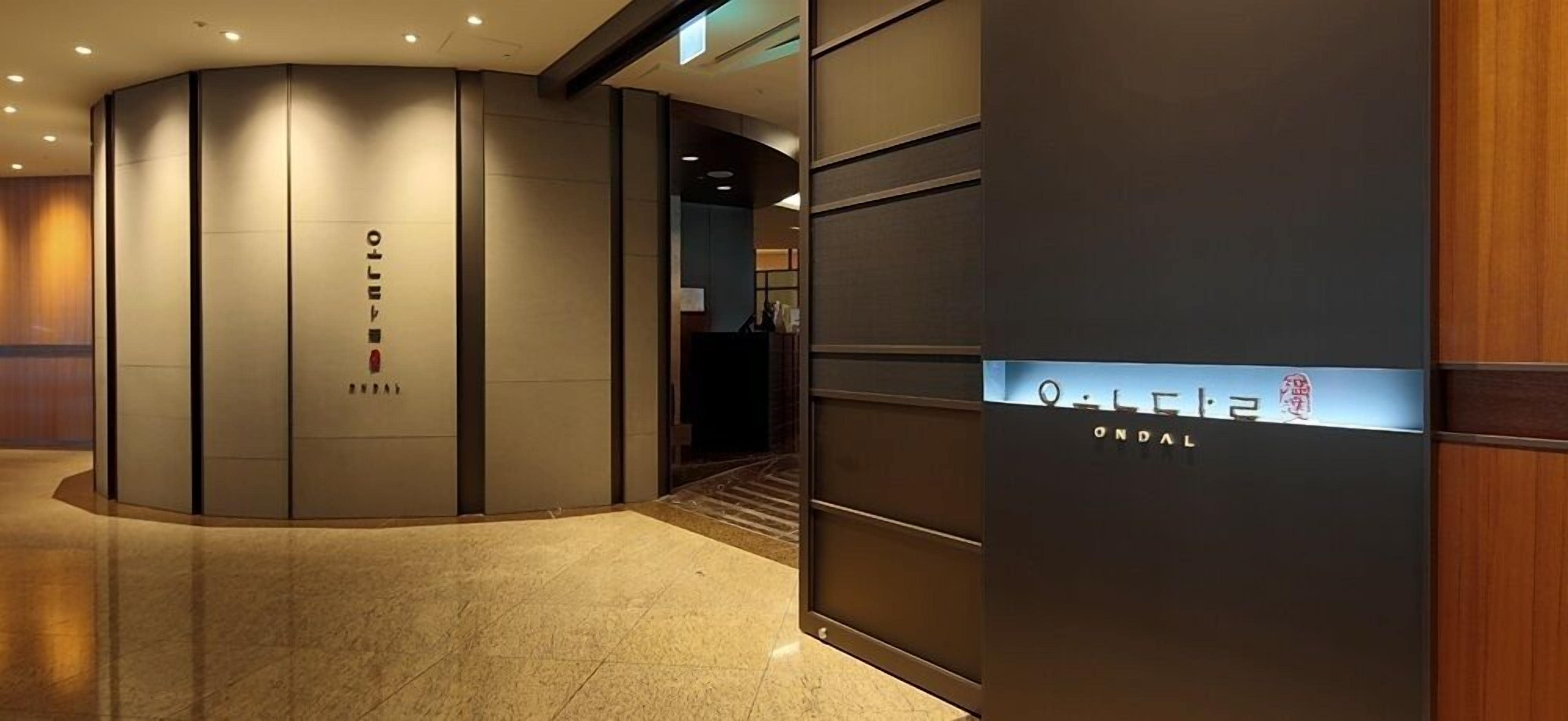 A modern hotel lobby with a curved reception desk, a digital display, and a staircase leading to the upper floors.