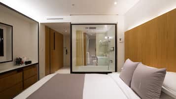 Habitación de hotel moderna con una cama grande, un armario de madera, un box de ducha de vidrio y un baño pequeño visible a través del cristal.