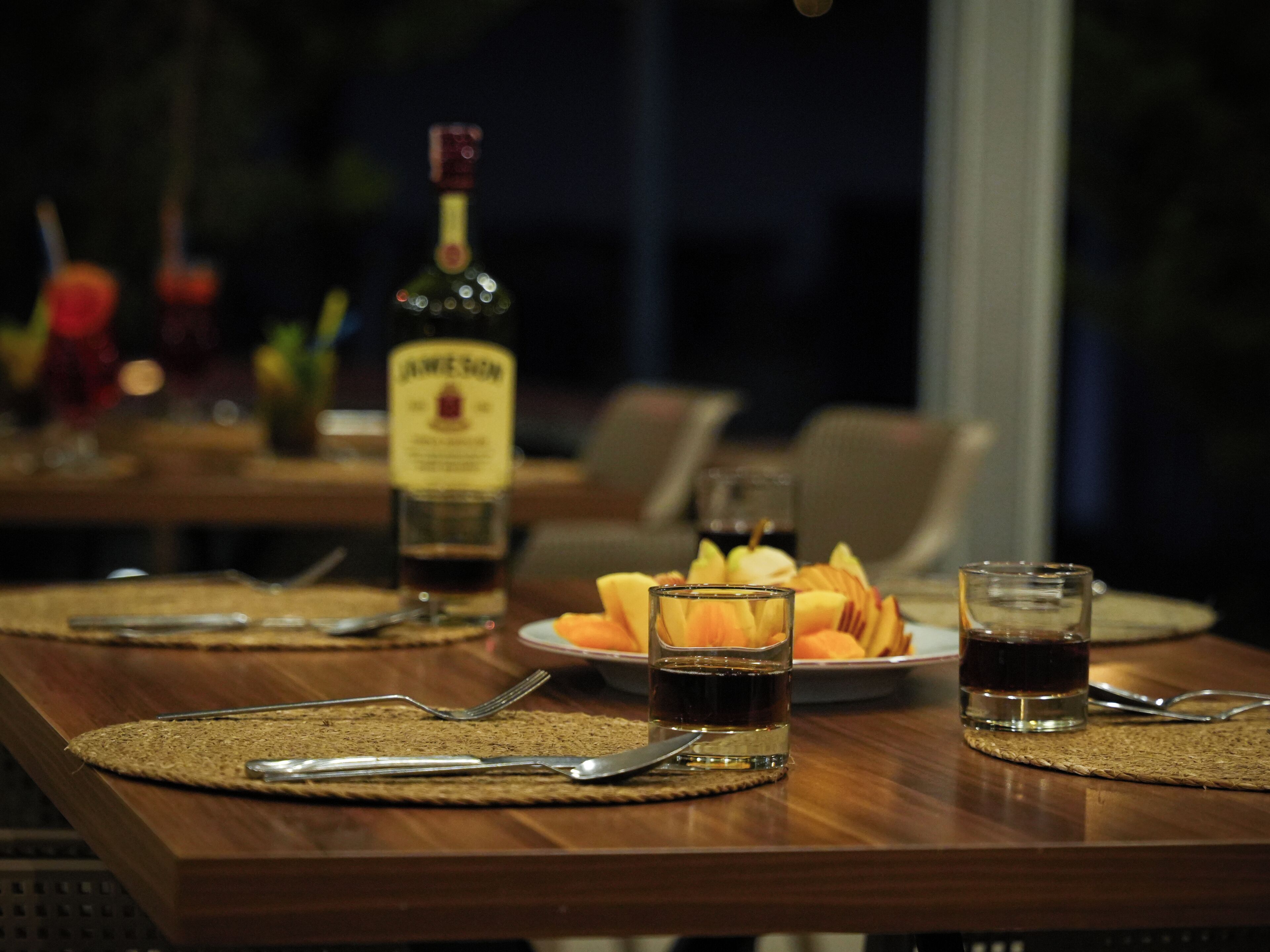 A dining table set with cutlery, plates, and glasses, with a bottle of liquor in the background.
