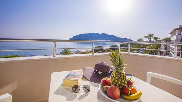 A balcony with a table set for a meal, a book, sunglasses, and a hat. A view of the sea and a distant island is visible. - Hawaii Suite Beach Hotel (Alanya)