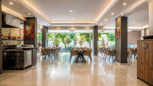 A modern restaurant with a central dining area, kitchen on the left, and large windows with a view of greenery. - Hawaii Suite Beach Hotel (Alanya)