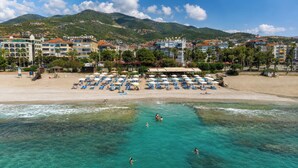 A beach with clear turquoise water, people swimming and sunbathing, and a row of beachside buildings with umbrellas. - Hawaii Suite Beach Hotel (Alanya)