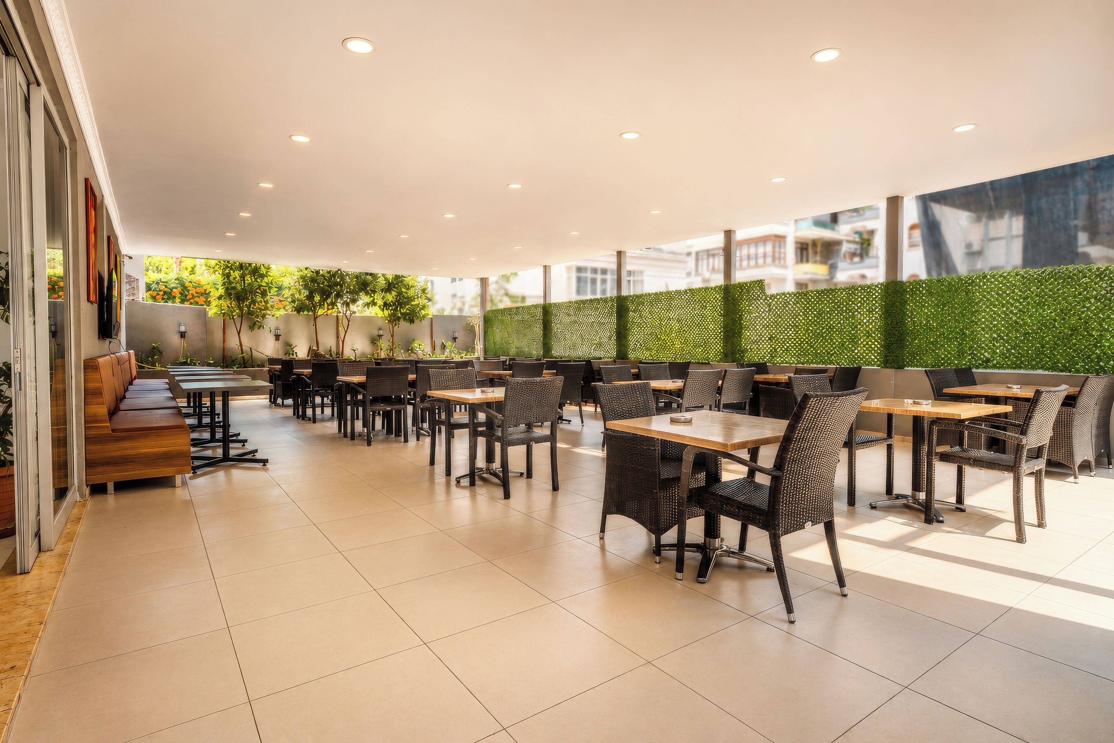 A spacious outdoor cafe with multiple tables and chairs, a green wall, and a view of the city.