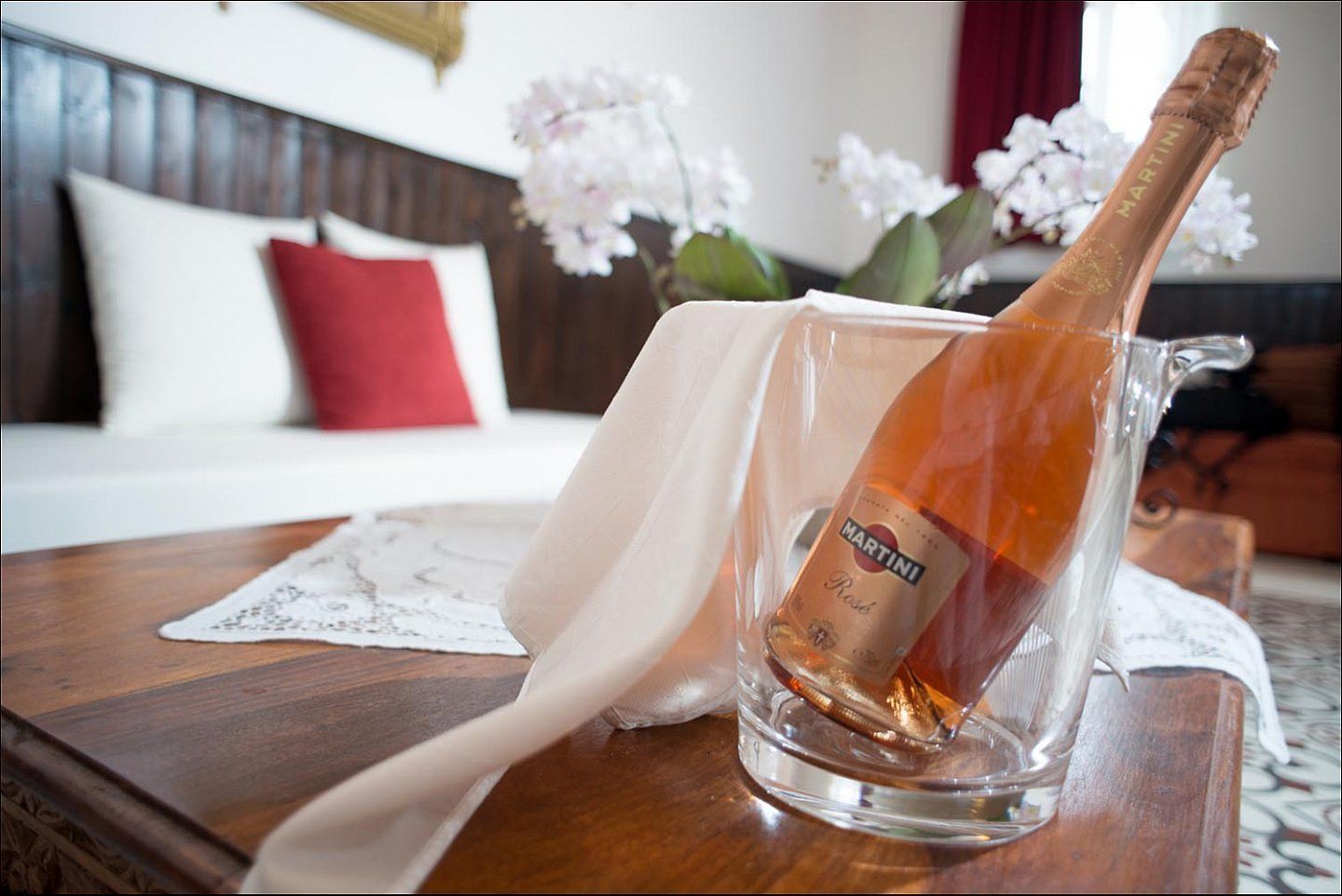 Een fles Martini Rosé champagne in een glazen karaf op een houten tafel met een witte tafelkleed en kantbeschermer.