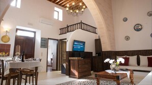 Amplia sala de estar con techo alto, vigas de madera, un candelabro, comedor con mesa puesta y un televisor sobre un mueble de madera.