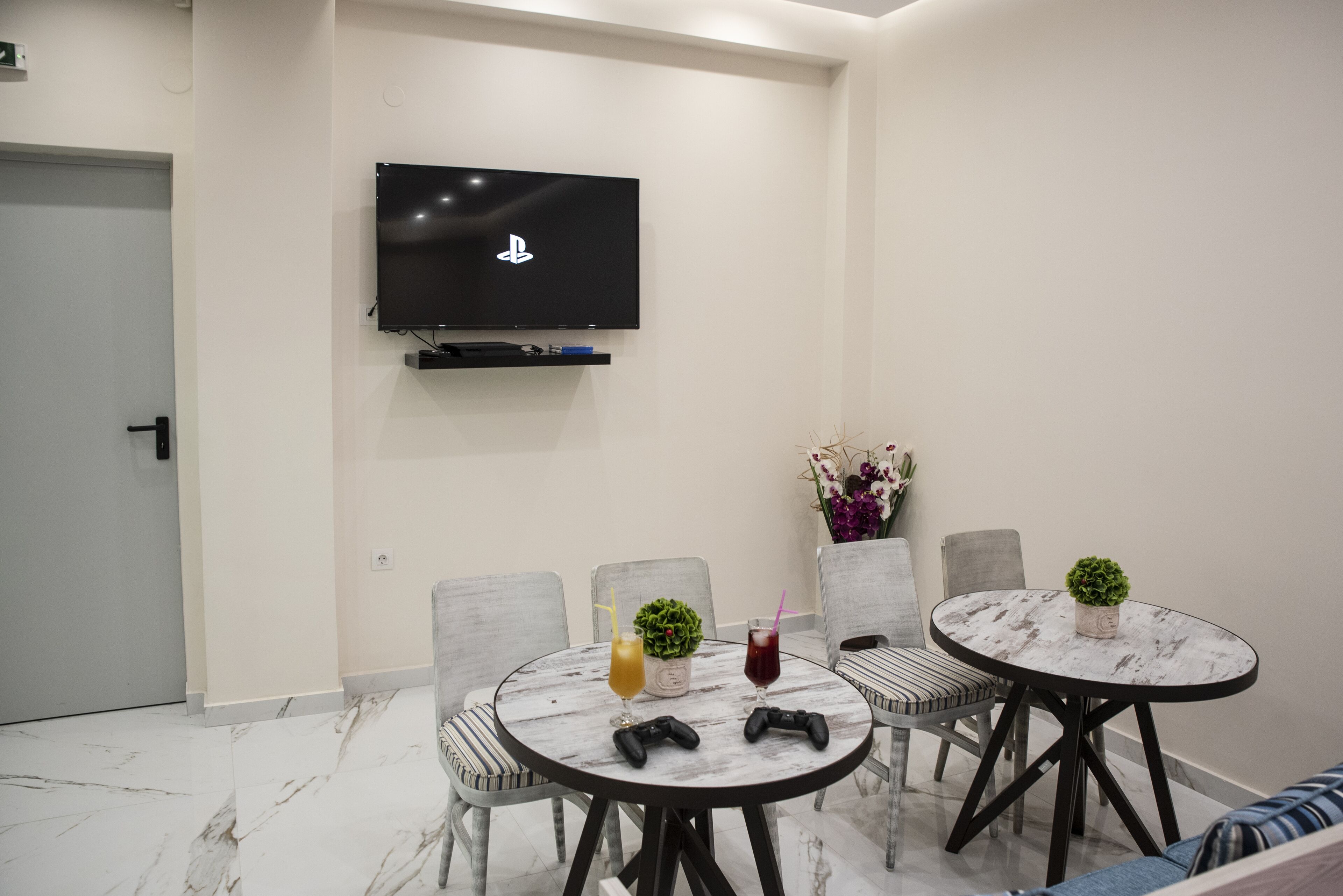 Una habitación moderna con un televisor de pantalla plana montado en la pared, una mesa redonda con sillas, dos mandos de videojuegos y dos bebidas sobre la mesa.