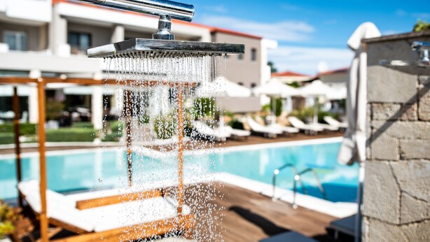 A modern outdoor pool area with a water feature, lounge chairs, and a clear blue sky. - Lagaria Apartments (Kassandra)