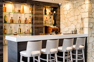 Uma área de bar com bancada branca, várias prateleiras com garrafas e três banquetas.