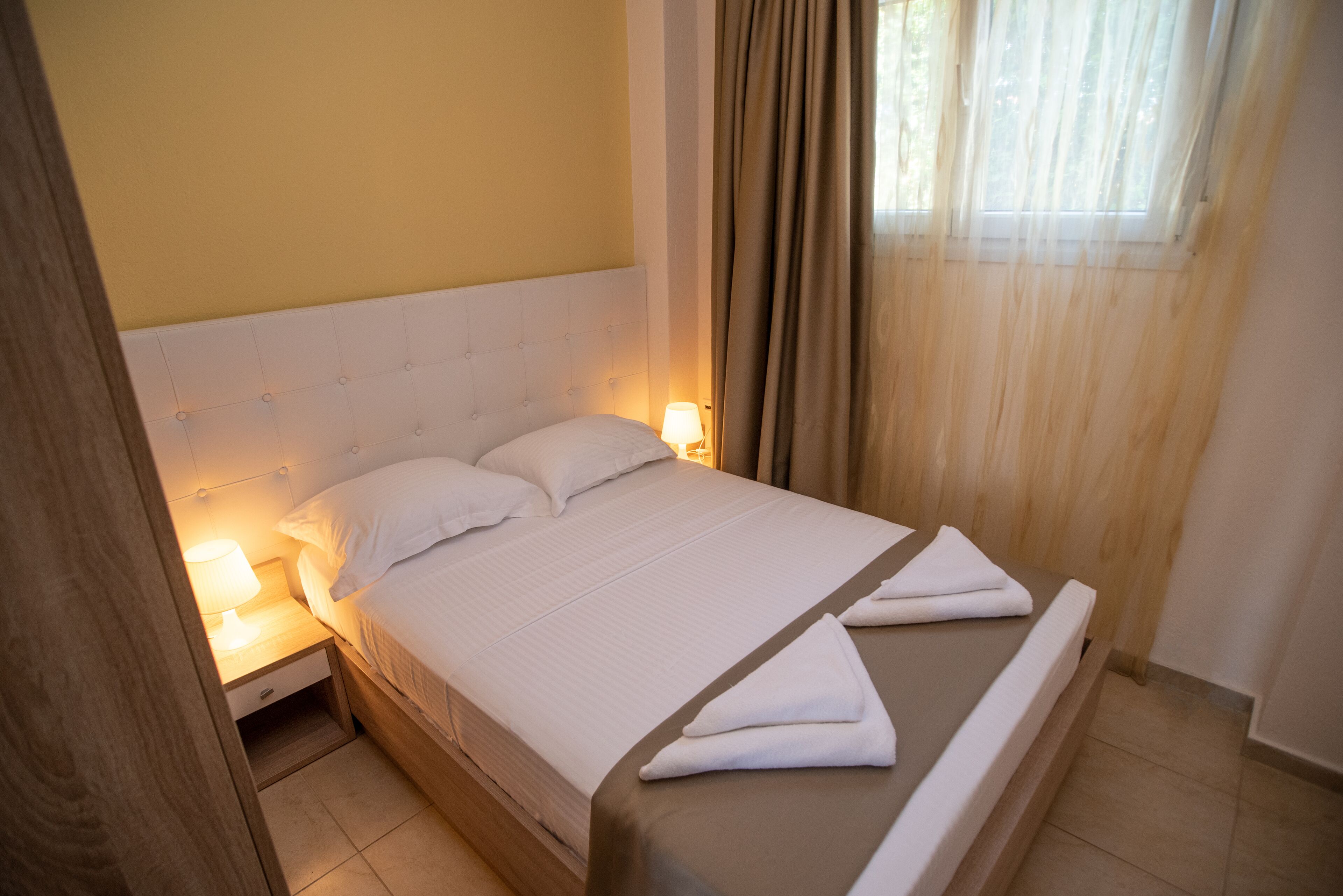 Una cama bien tendida con sábanas blancas y un cabecero marrón, una lámpara de noche y una ventana con cortinas.