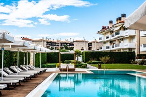 A swimming pool with lounge chairs and umbrellas, surrounded by buildings with balconies and a well-maintained garden. - Lagaria Apartments (Kassandra)