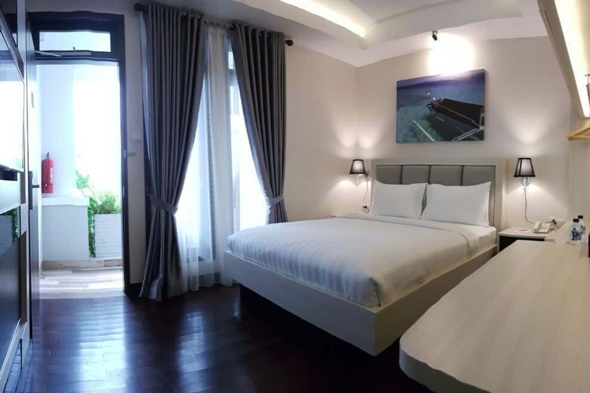 Una habitación de hotel con una cama grande, mesitas de noche, un escritorio, una silla, una ventana con cortinas, un cuadro en la pared y un extintor de incendios en la esquina.