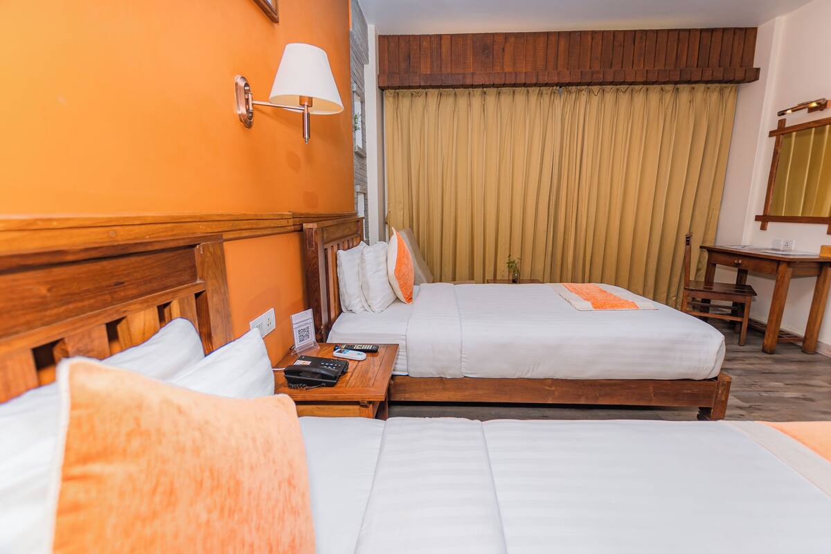 A hotel room with two beds, a wooden headboard, a bedside table with a lamp, and a window with curtains.
