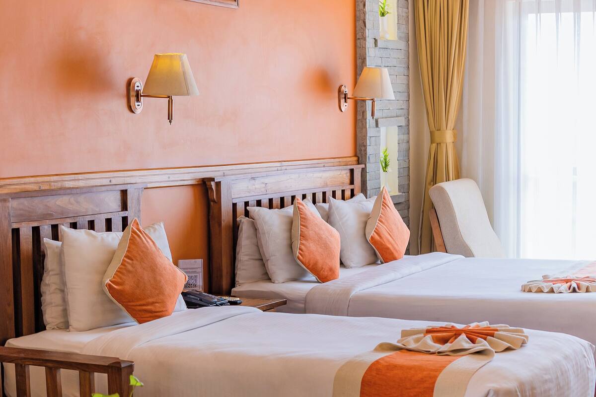 A hotel room with two beds, a wooden headboard, a small table with a vase, and a painting on the wall.