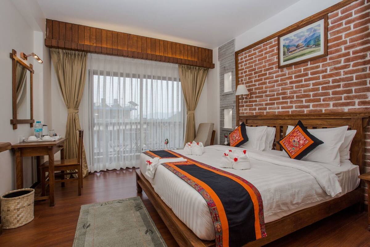 A hotel room with a wooden bed, a desk with a chair, a large window with curtains, and a brick wall with a painting.
