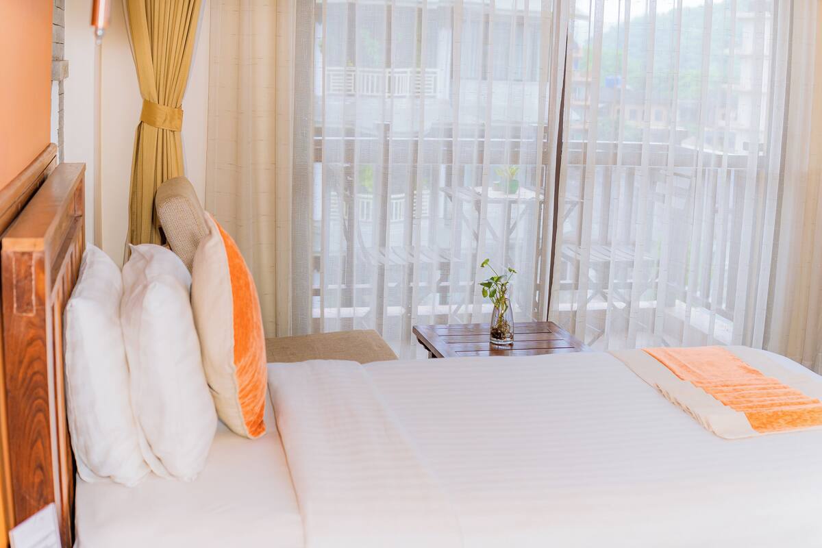 A hotel room with a bed, pillows, a small table with a vase, and a view through sheer curtains.