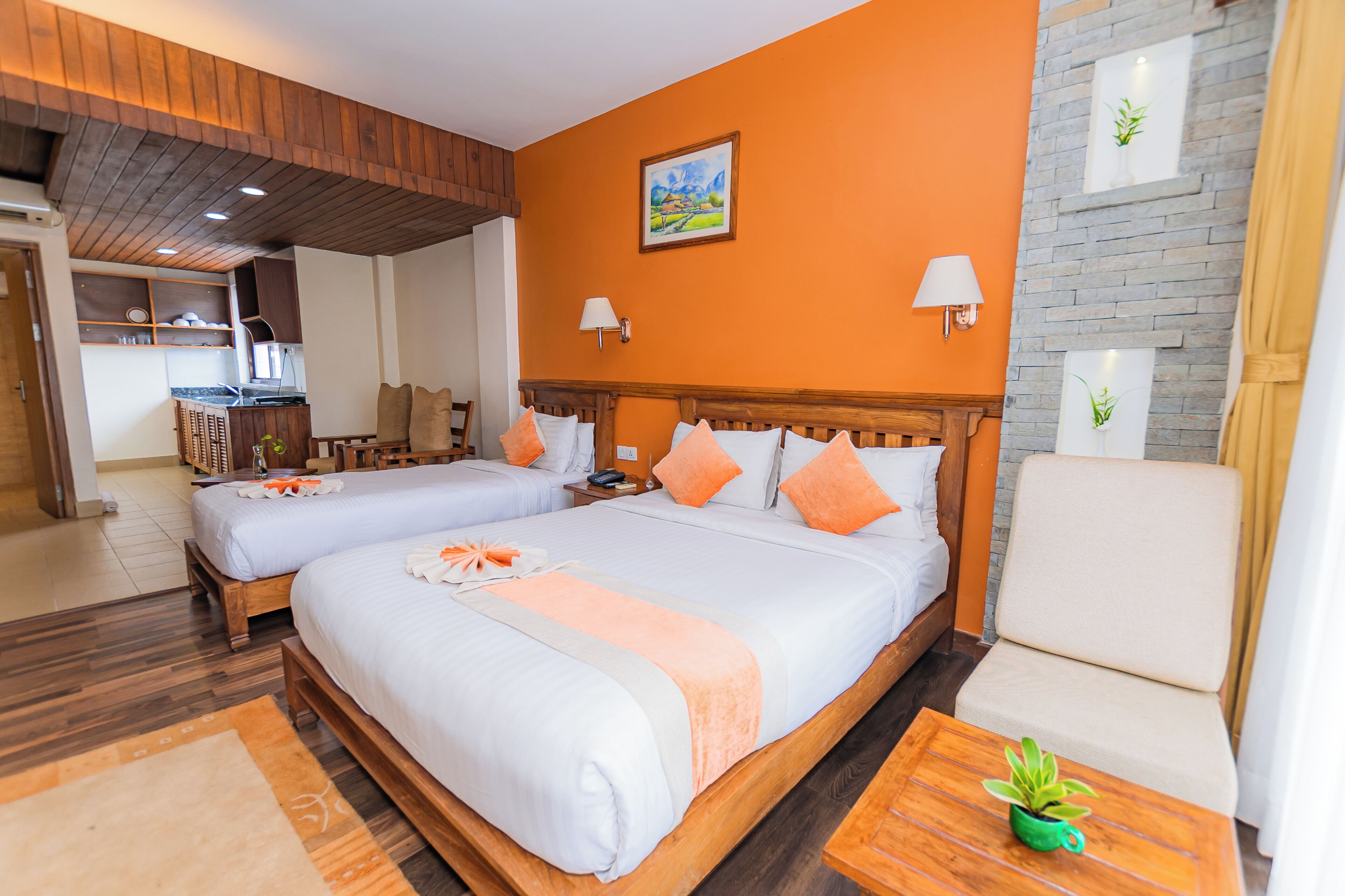 A hotel room with two beds, a wooden headboard, a green plant on a side table, and a view of the kitchen area.