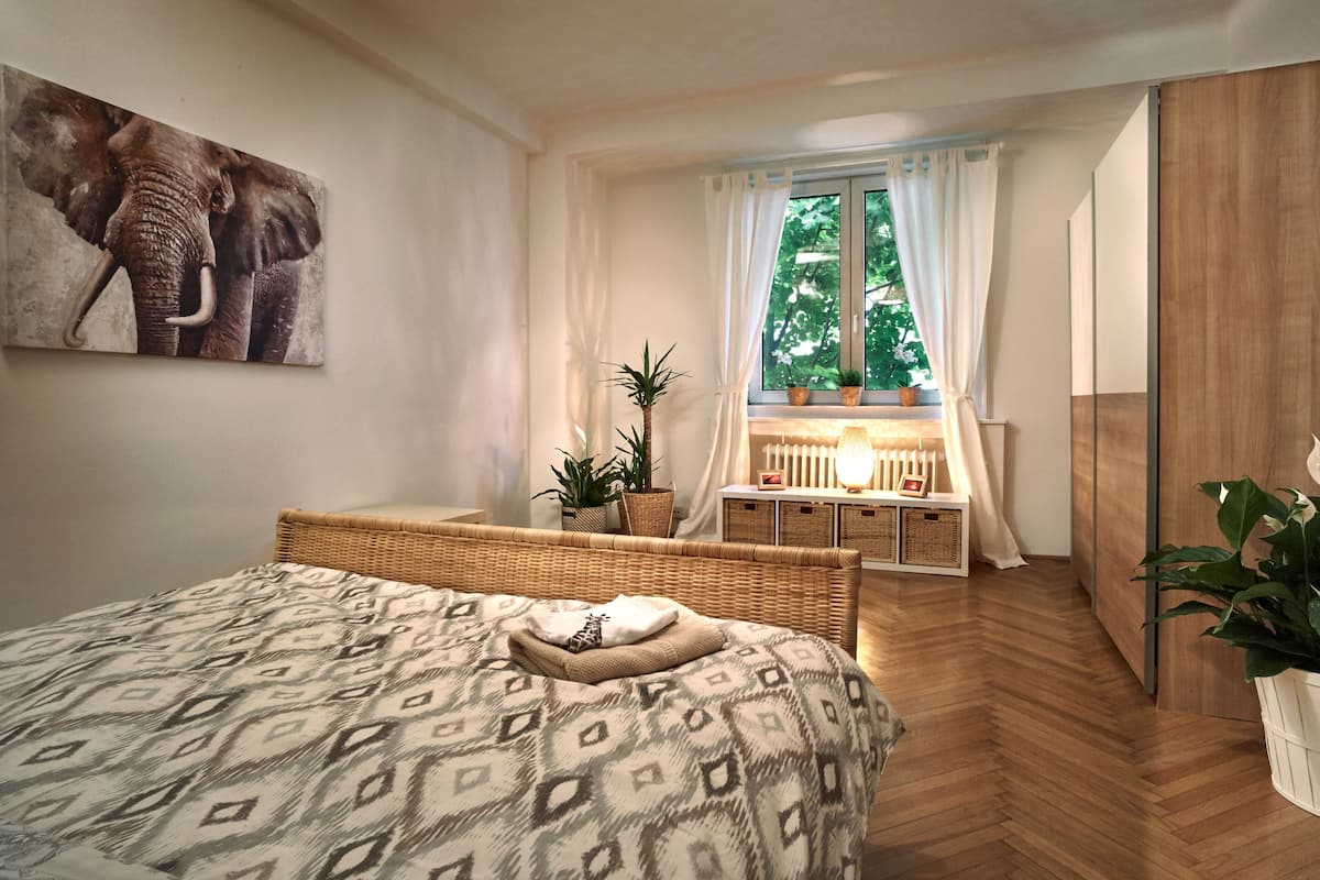 A bedroom with a patterned bedspread, a wicker headboard, a window with curtains, a bedside table with a lamp, and a potted plant.
