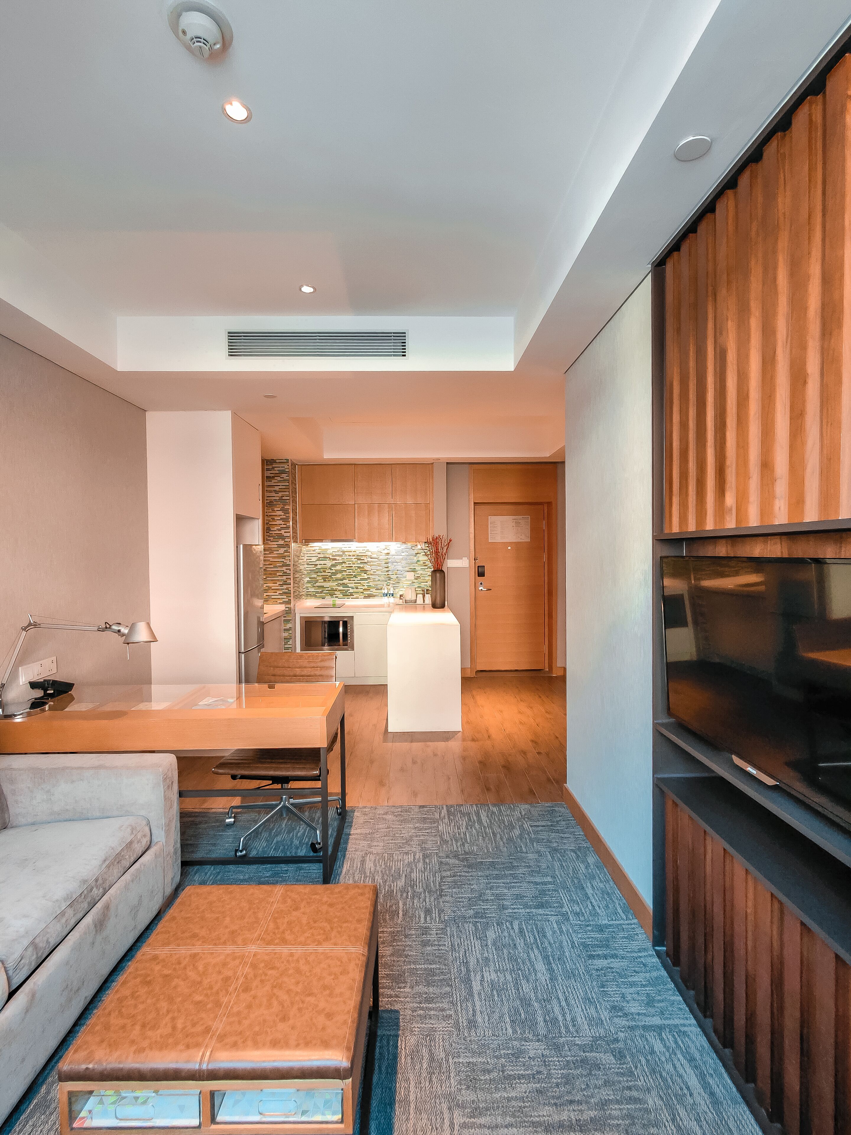 A modern living room with a sofa, a coffee table, and a TV. There is a kitchen area with a microwave and a refrigerator.