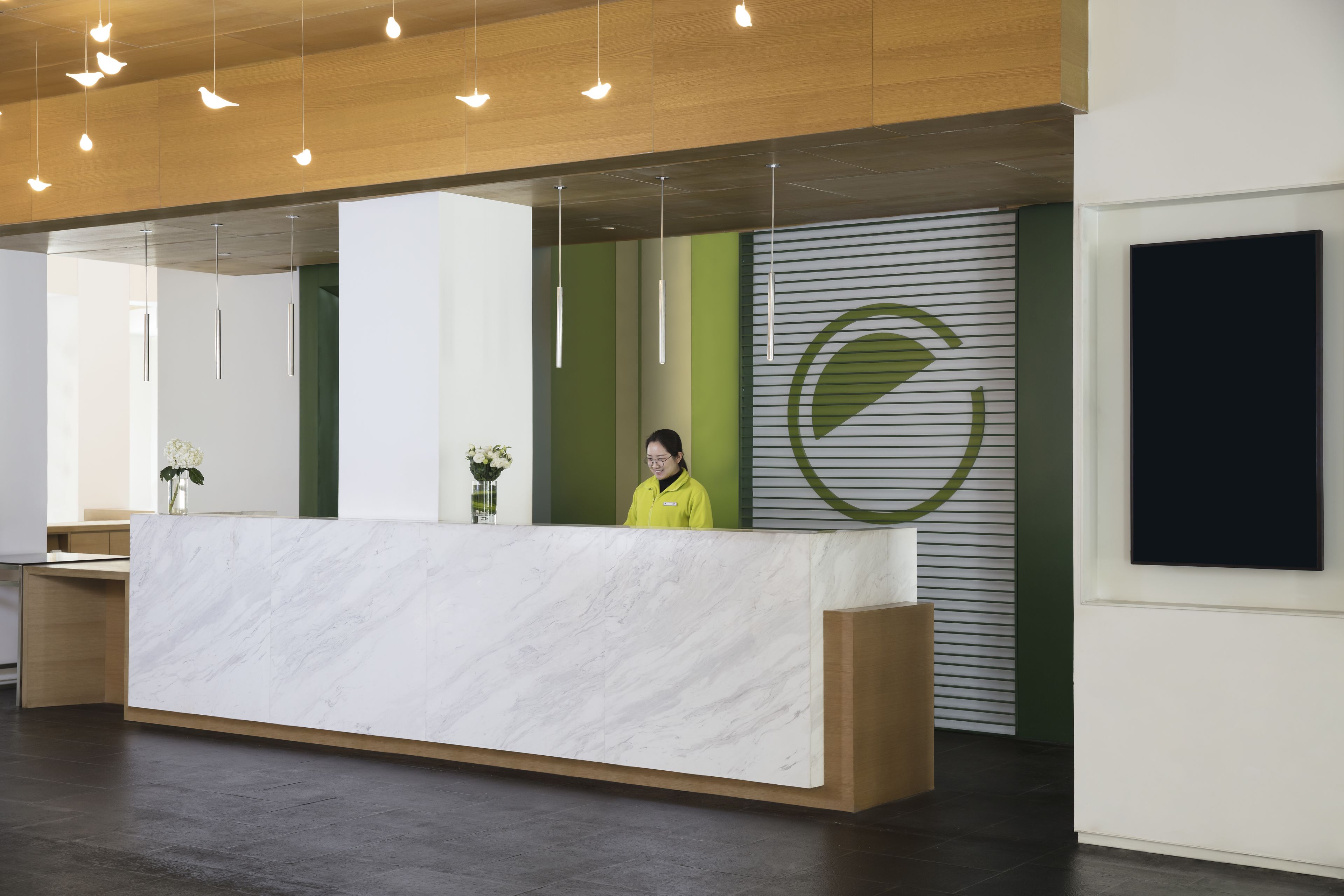 Una recepción con mostrador de mármol, una mujer de pie detrás y una pared verde y blanca con un logotipo circular.