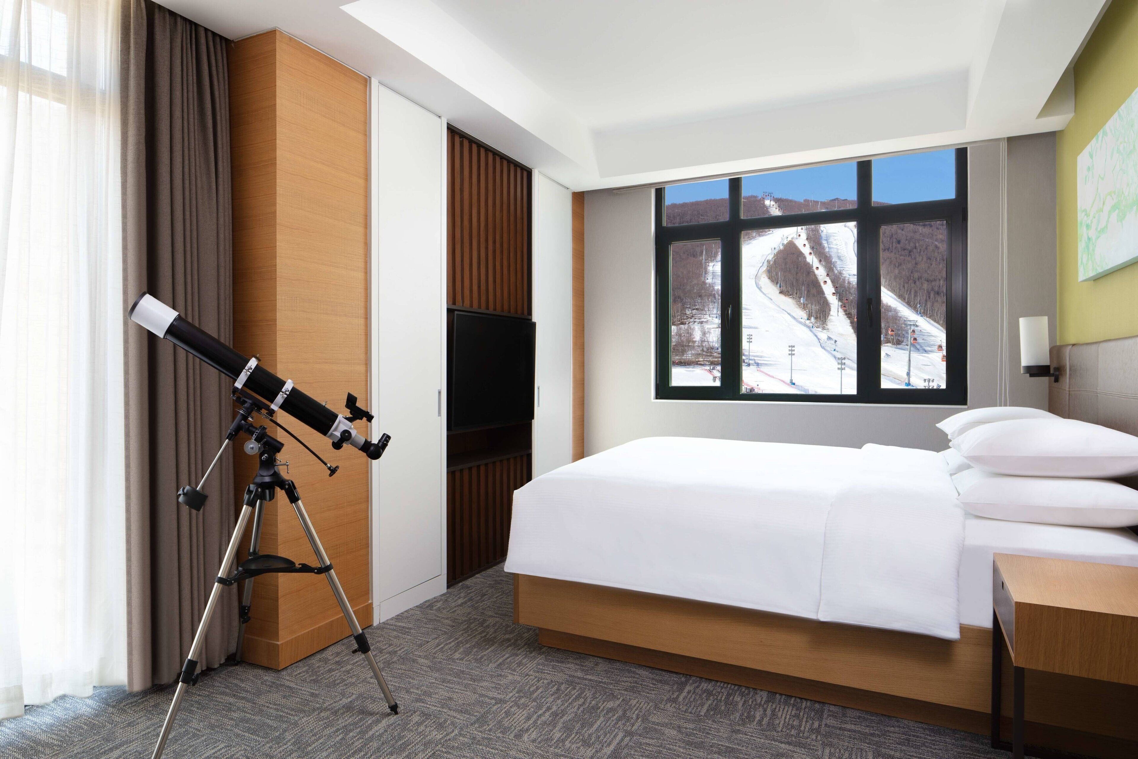 A hotel room with a bed, a television, a telescope, and a window with a view of a snowy landscape.