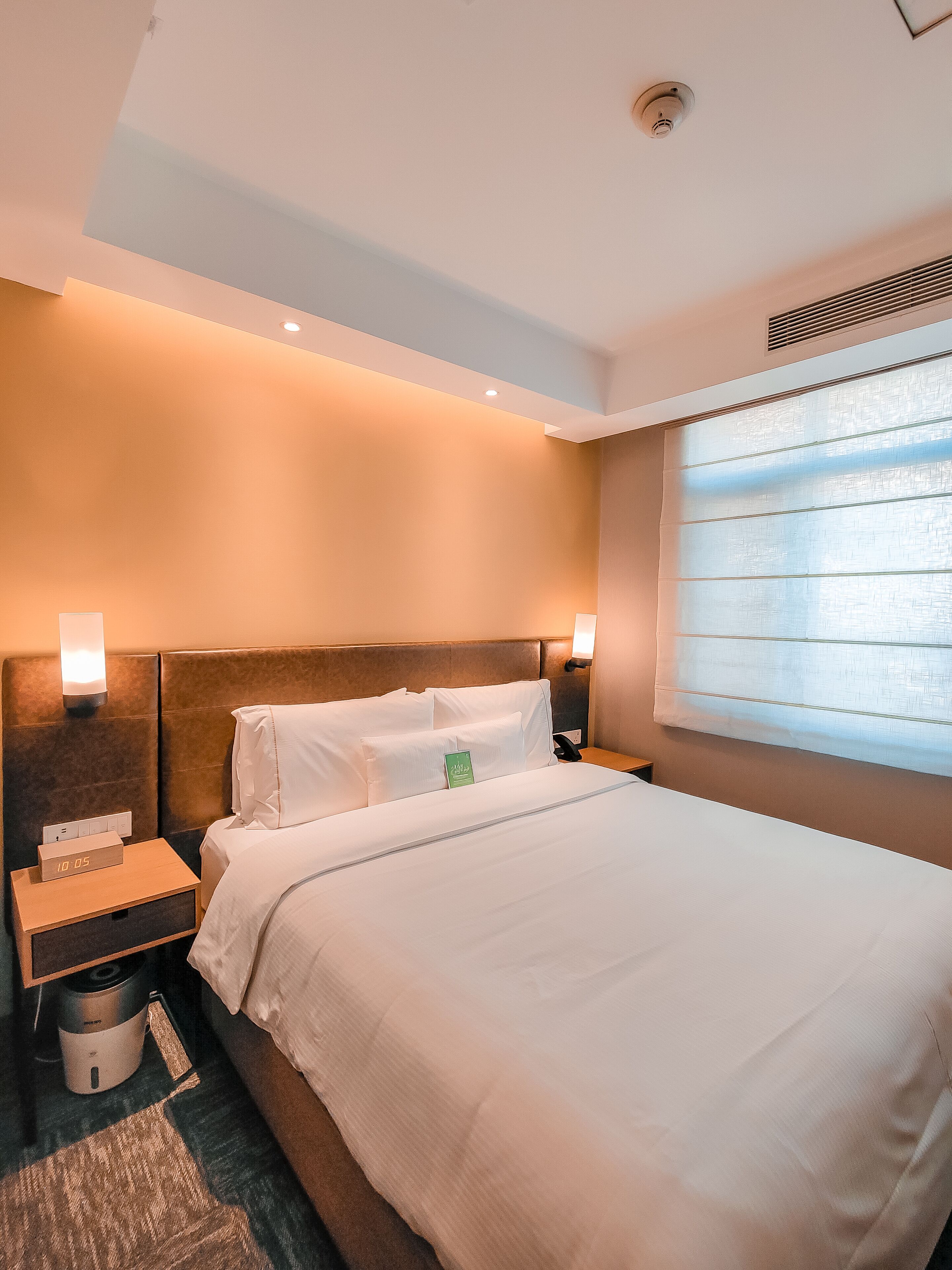 Une chambre d’hôtel avec un grand lit, une table de chevet dotée d’une horloge numérique, une autre table de chevet avec une lampe, et une fenêtre avec des stores.