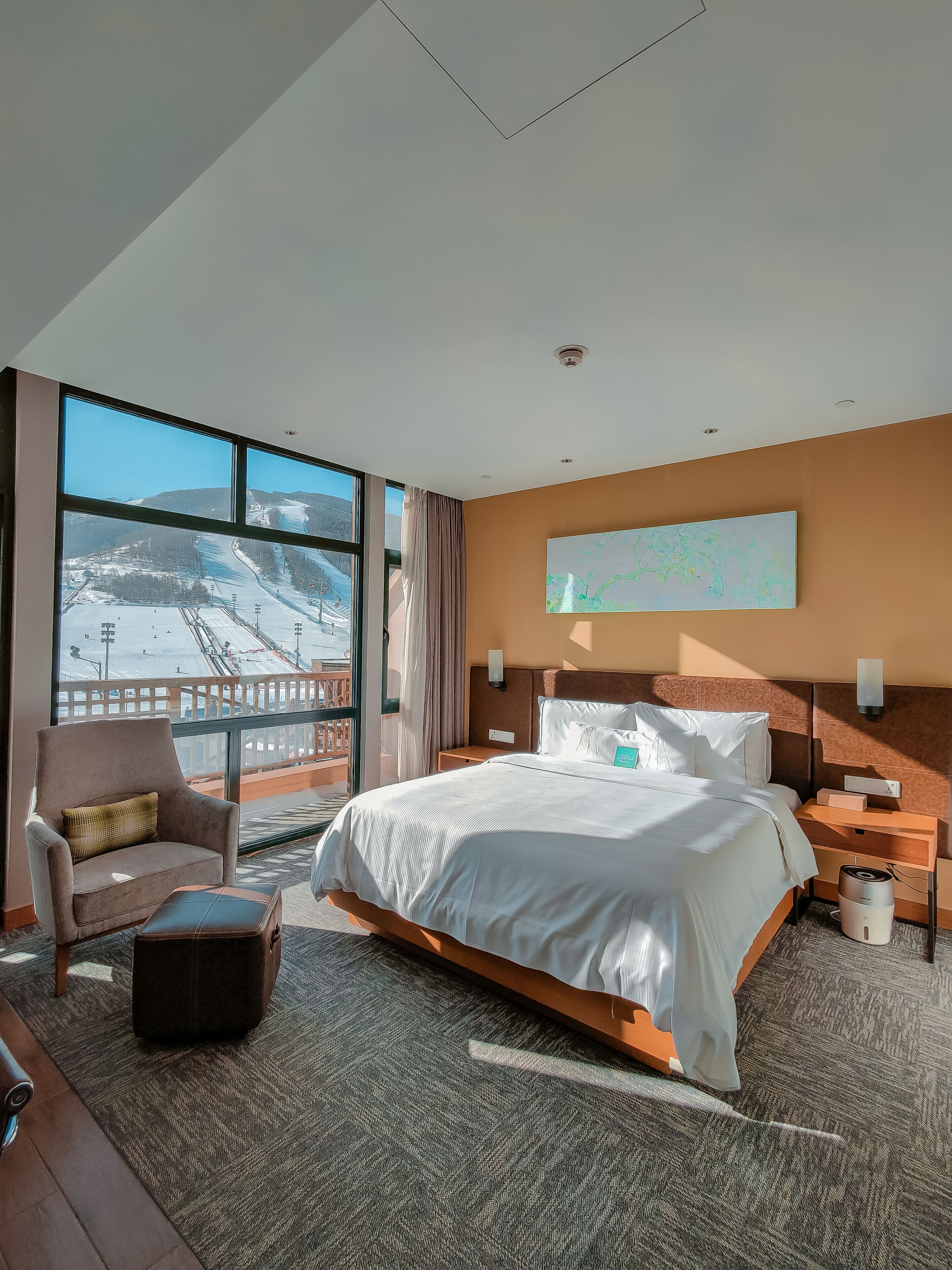 Una habitación de hotel con una cama grande, una silla, una mesita y vistas a un paisaje nevado a través de amplios ventanales.