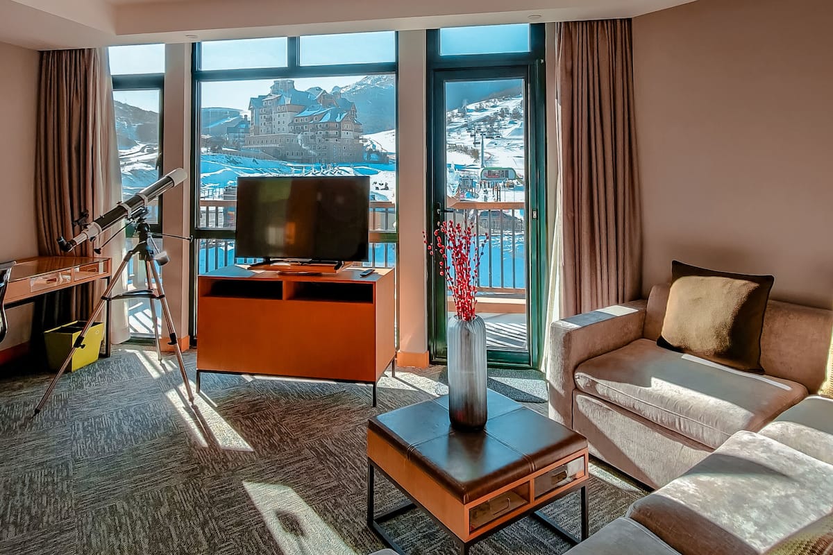 A modern hotel room with a large window offering a scenic mountain view, a comfortable sofa, a television, and a telescope.