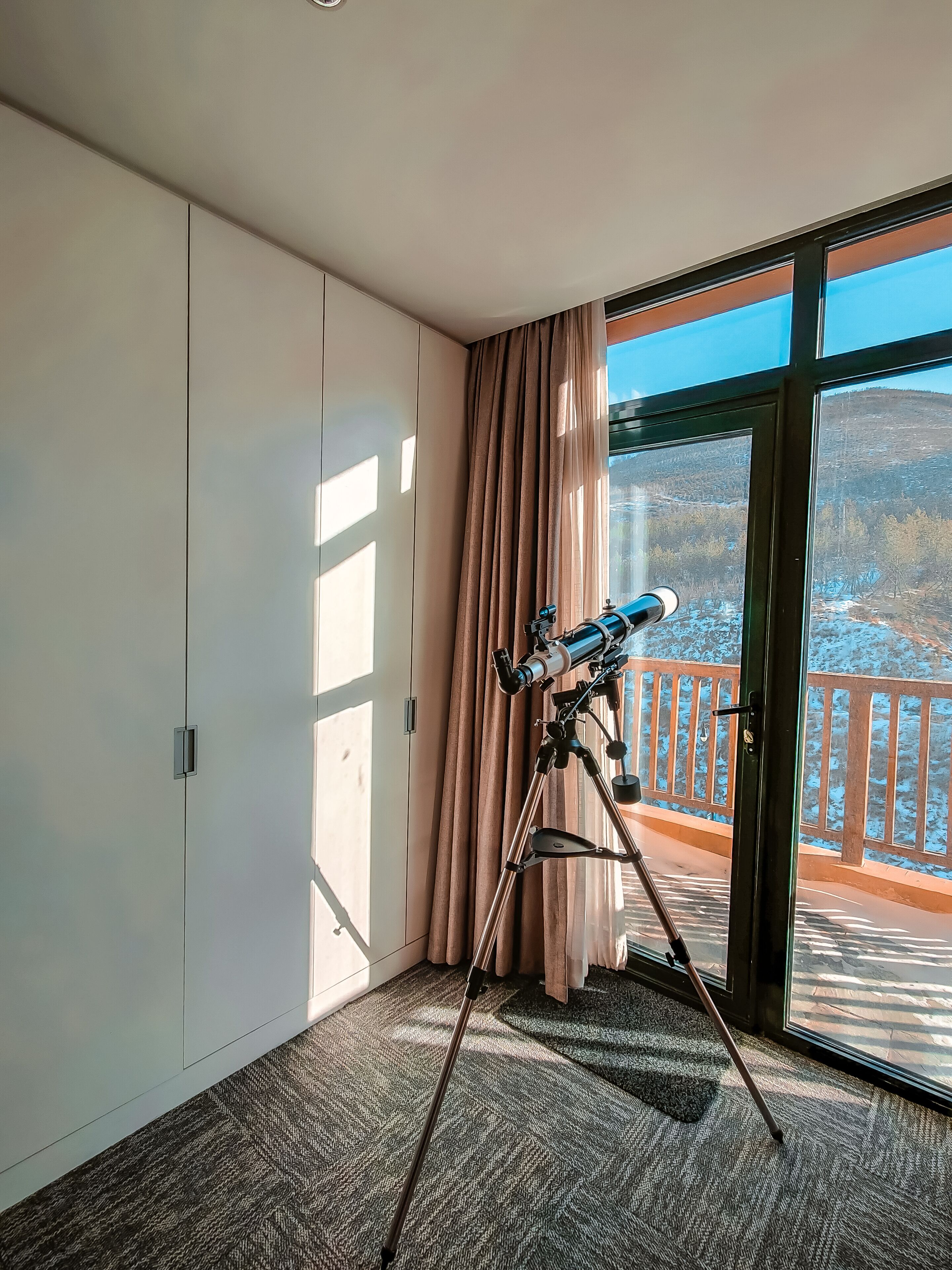 A room with a telescope on a tripod, a sliding glass door leading to a balcony, and a carpeted floor.