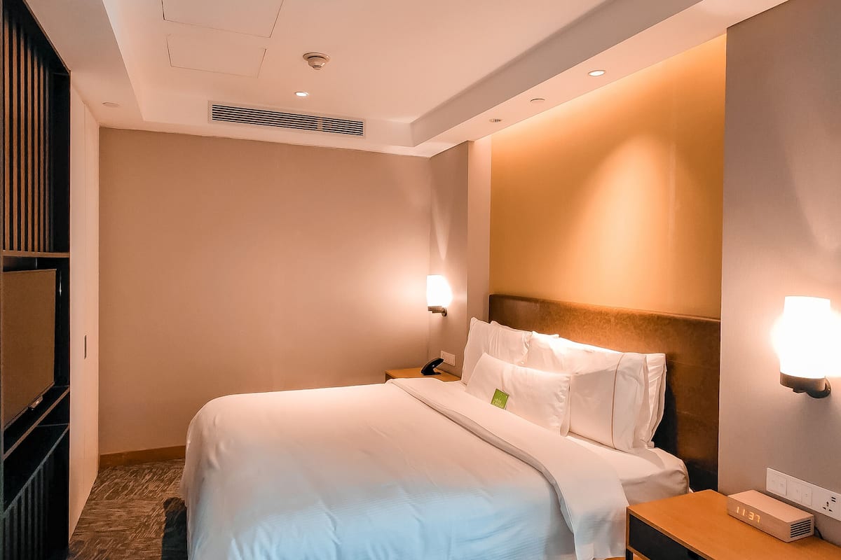 A hotel room with a neatly made bed, a bedside table with a lamp, a wall-mounted heater, and a small bedside clock.