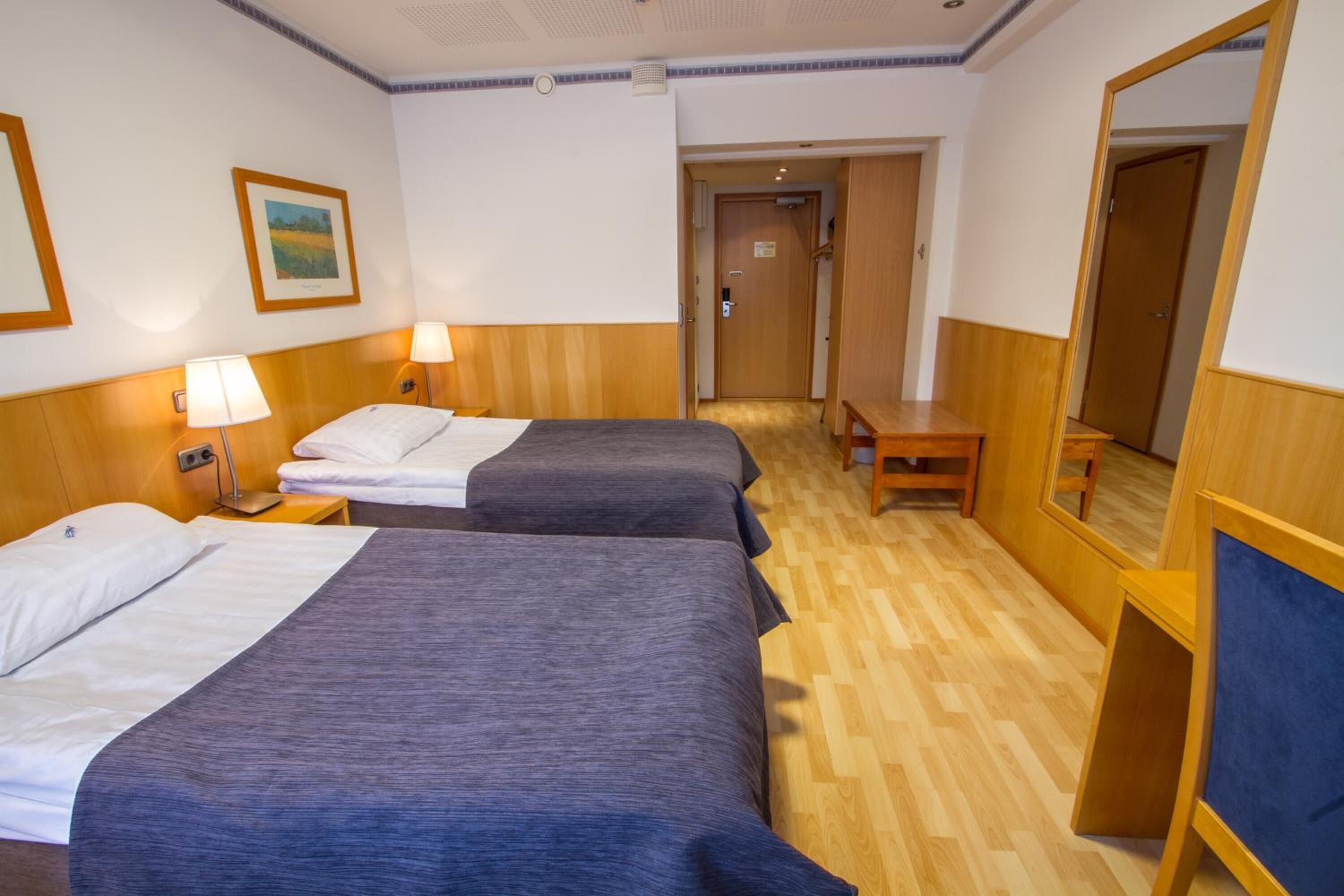 A hotel room with two beds, wooden flooring, a mirror, and a small table.