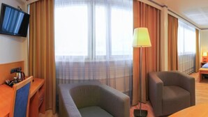 Quarto de hotel com cama, duas poltronas, escrivaninha com luminária, televisão e banheiro visível por uma porta aberta.