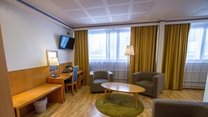 Quarto de hotel com uma escrivaninha de madeira, duas poltronas, um sofá, uma TV fixada na parede e janelas amplas com cortinas.