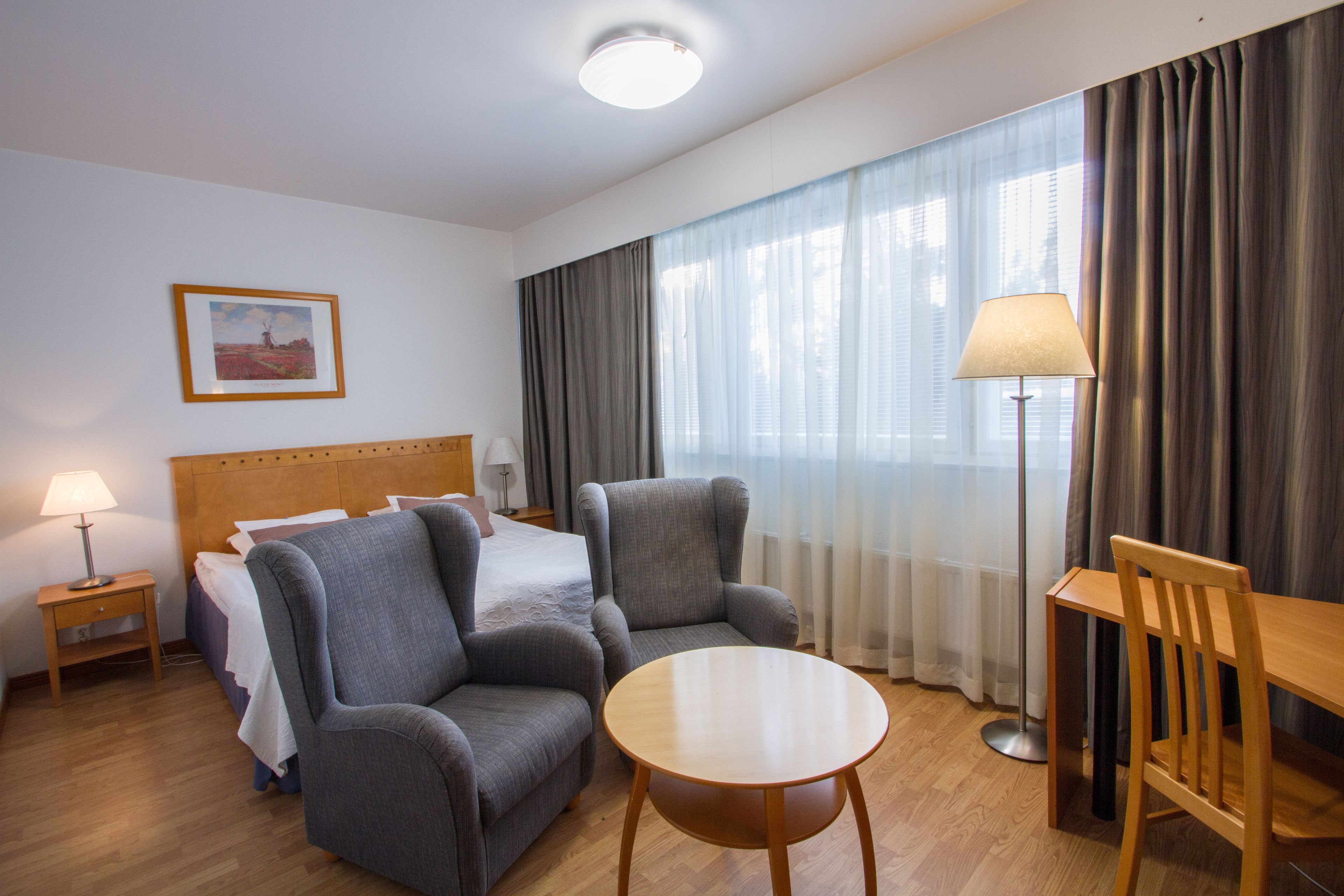 Hotellihuone, jossa on sänky, kaksi nojatuolia, pyöreä pöytä, lattiavalaisin ja taulu seinällä.