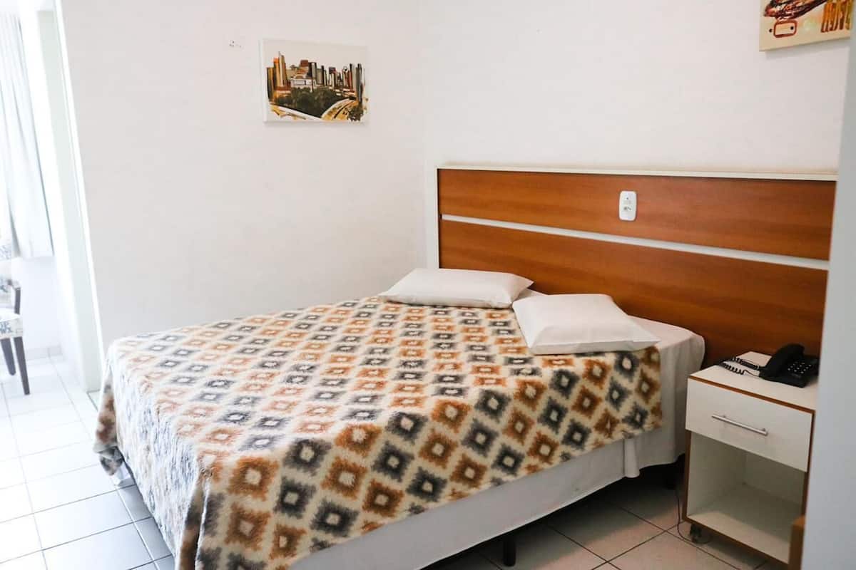 Una cama bien hecha con una colcha estampada, un cabecero de madera, una mesita de noche con un teléfono y un cajón pequeño.