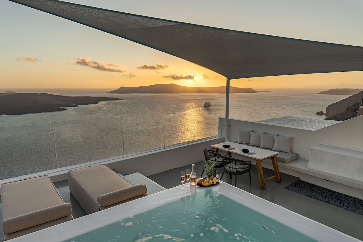 Una terraza moderna al aire libre con piscina, mobiliario y una mesa lista para dos personas, con vista a un paisaje costero al atardecer.
