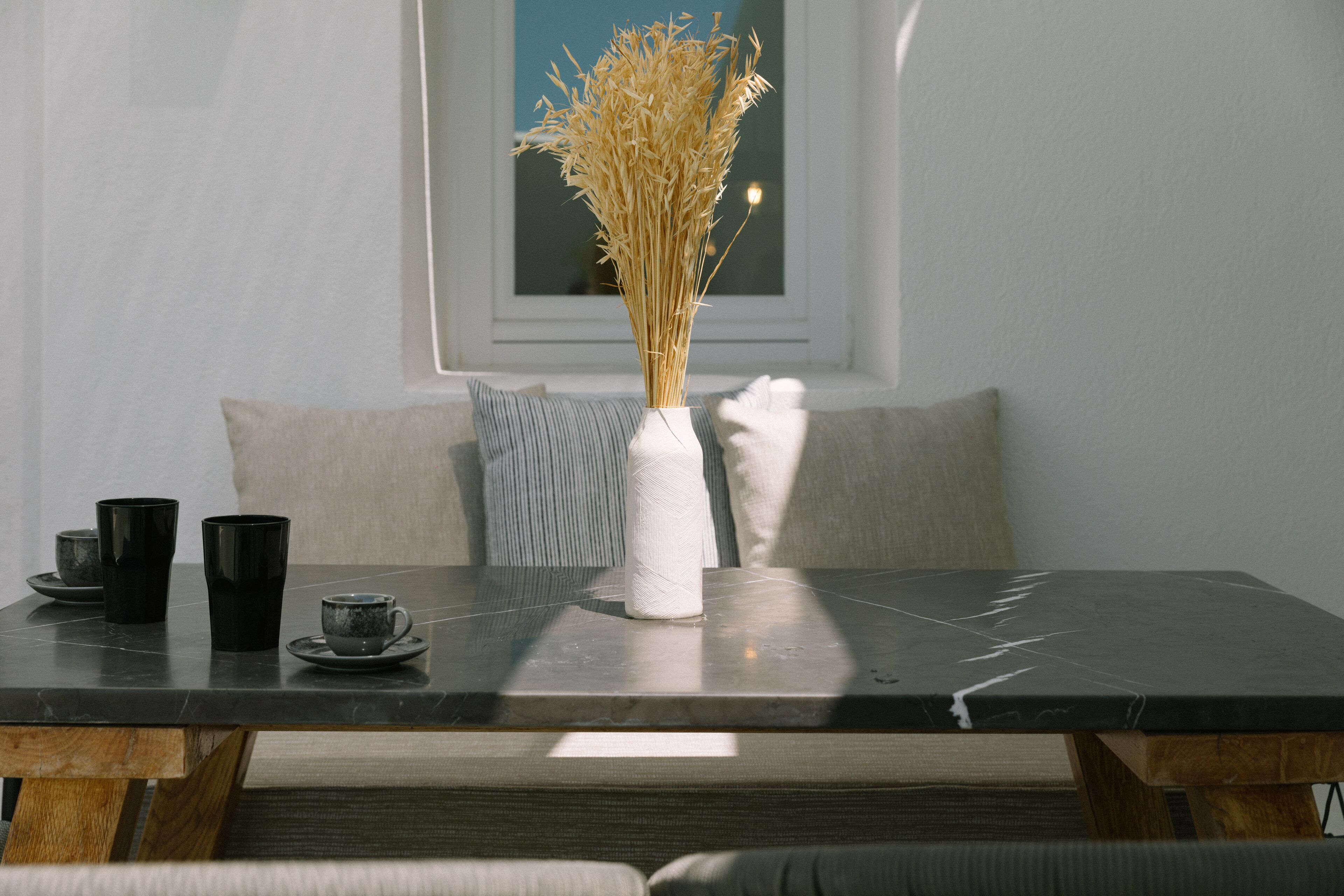 A dining table with a vase of dried plants, black cups, and a wooden bench.