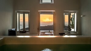 A room with a view of the sea at sunset, a small table with drinks, and a pool in the foreground. - Thera Villas (Santorini)