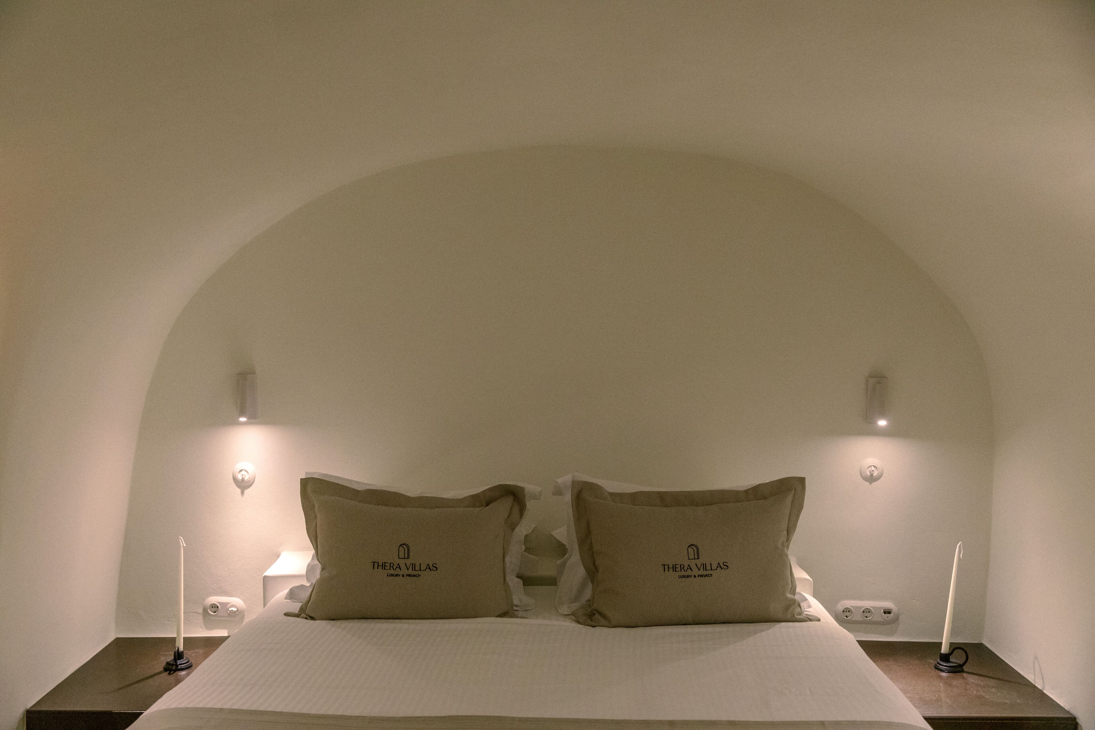 A neatly made bed with two pillows, a bedside table with a lamp, and a wall-mounted light fixture.