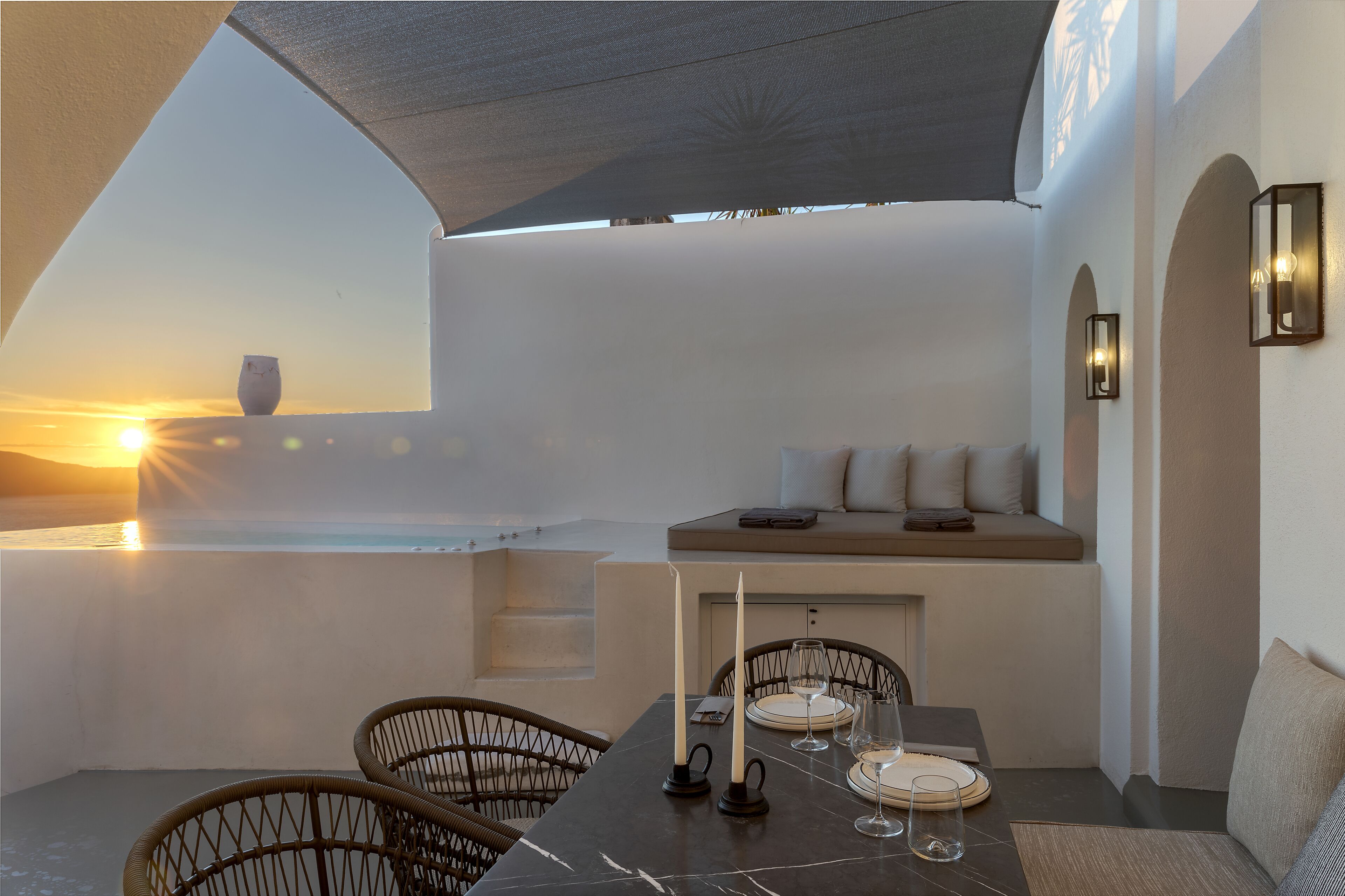 A dining area with a table set for two, a view of the sunset, and a seating area with cushions.
