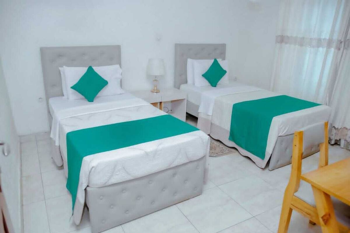 Habitación de hotel con dos camas, cada una con cabecero blanco y colcha verde. Hay una silla de madera y una mesita en la habitación.