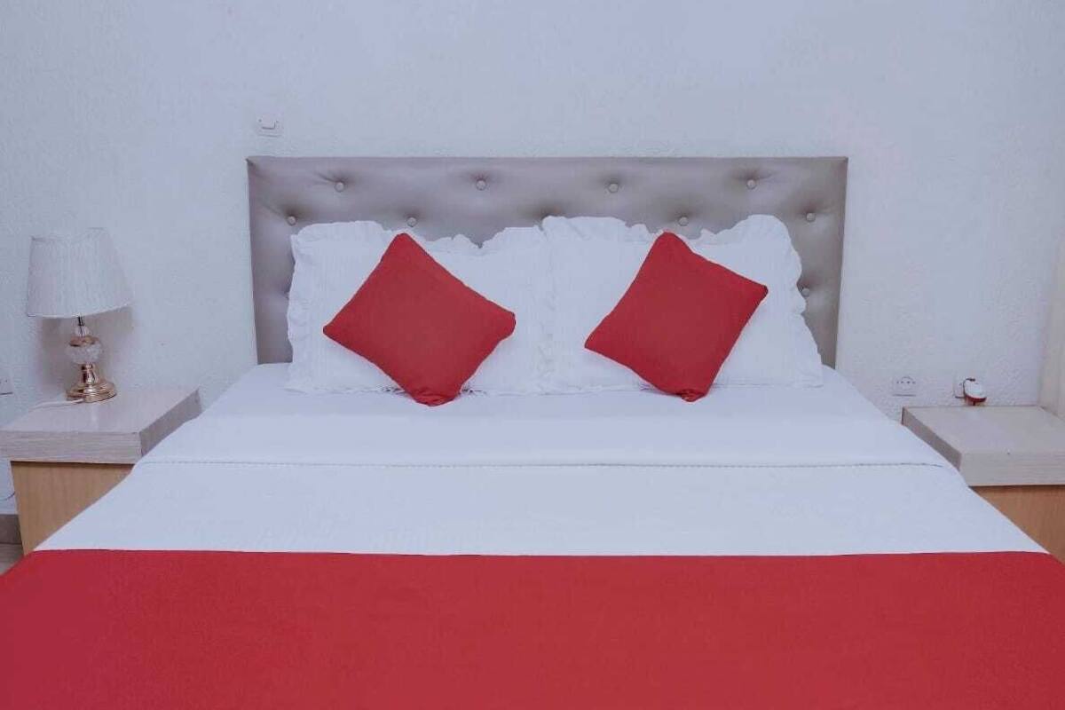 Una cama bien tendida con una colcha blanca y roja, dos almohadas rojas y un cabecero capitonado.